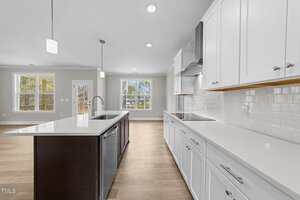 A modern kitchen with white cabinets, white countertops, a dark island with a sink, pendant lighting, and light wood floors. Large windows and a glass door provide natural light and a view of the backyard.
