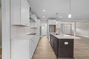 A modern kitchen with white cabinets, light countertops, a central island with a dark base, stainless steel appliances, a tile backsplash, and light wood floors, opening into a spacious living area with large windows.