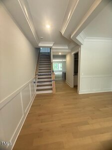 A hallway with light wood flooring, white wainscoting and walls, a wooden staircase with a light railing on the left, and an open view to a bright room with windows in the background.