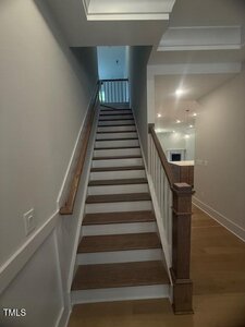 A straight wooden staircase with white risers and a wooden handrail leads upward in a modern, well-lit home interior with light-colored walls and flooring.