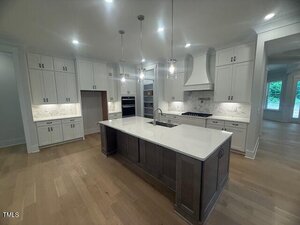 Modern kitchen with white cabinets, stainless steel appliances, a center island with a dark wood base and white countertop, pendant lights above, and light wood flooring. The kitchen opens into another room with large windows.