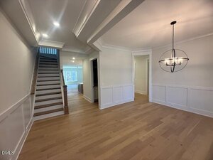 A modern, empty interior with light wood floors, white walls with wainscoting, a staircase on the left, a dining area with a round chandelier, and an open view into another room in the background.