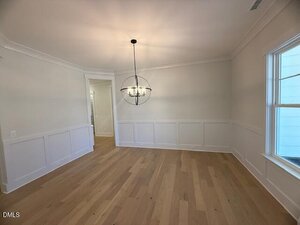 A dining room with light wood floors, white wainscoting, a large window, and a modern spherical chandelier hanging from the ceiling. The walls are painted light beige and the room opens into a hallway.
