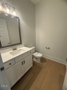 A small bathroom with light wood flooring, a white vanity with a sink and silver faucet, a wall-mounted mirror with lights above, and a toilet with a closed lid. The walls are painted light gray.