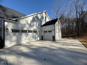 A white three-car garage with decorative windows and black lantern-style lights on the exterior, adjacent to a wooded area under a clear blue sky. The driveway is wide and made of smooth concrete.
