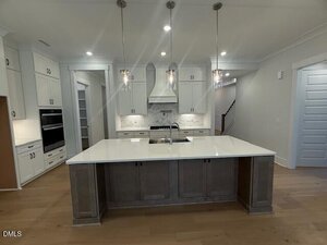 A modern kitchen with a large island featuring a sink, pendant lighting, white cabinetry, built-in oven, gas stove, white tile backsplash, light wood floors, and a staircase visible in the background.
