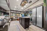 Modern RV interior at 115 Squaw Drive, Louisburg, featuring a kitchen island, double-door refrigerator, built-in microwave, sofa, recliner chairs, large windows with outdoor views, overhead cabinets, wood-style flooring, and sleek lighting fixtures.