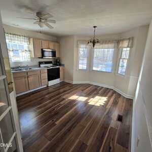 A kitchen with light wood cabinets, a white stove, and a black dishwasher. There is a bay window area with a hanging light fixture and wood flooring throughout, with sunlight streaming in from the windows.