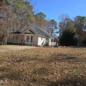 A white single-story house with a front porch sits on a large yard covered in dry leaves. Children's toys and play equipment are scattered near the house. There are trees with sparse leaves in the background under a clear blue sky.