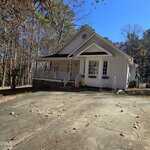A single-story white house with a front porch and gable roof sits next to a wooded area at 122 Buckskin Drive, Louisburg. The wide, cracked driveway leads to a yard where toys and a small American flag are visible near the porch.