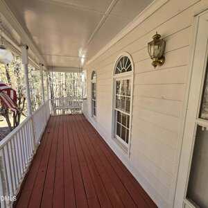 A covered porch with red wooden flooring, white railing, and beige siding. Two large arched windows and a brass wall-mounted light fixture are visible. An American flag hangs on the left side. Trees can be seen in the background.