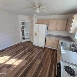 A kitchen with wood flooring, light-colored cabinets, a white refrigerator, a white stove, a stainless steel sink, and a ceiling fan. A glass-paneled door is on the left side of the room.