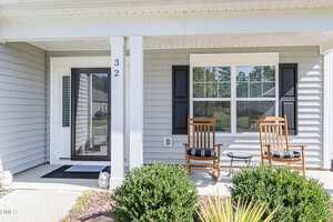 A covered front porch with two wooden rocking chairs, a small round table, and black shutters. The house has light gray siding, white trim, and the number 32 is displayed on a porch post. Shrubs line the edge of the porch.