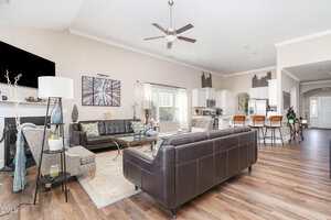 Open-concept living space with dark leather couches, a coffee table, and accent chairs. The kitchen features white cabinets, stainless steel appliances, and a breakfast bar with wooden stools. There is wood flooring throughout.