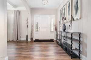 A modern entryway with wood flooring, a white front door, two narrow windows, contemporary decor on a black console table, tall vases, and framed artwork on a beige wall. Light fills the space.