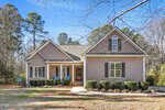Charming single-story house at 1511 Anterra Drive, Wake Forest, with tan siding, white trim, and a front porch. Features gabled roofs, large windows, a tidy yard with trimmed bushes and a curved walkway. Tall trees in the background.