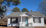 A small, single-story white house with a gray roof, black shutters, and a light blue front door sits at 51 Furney Pearce Road in Zebulon. The cozy front porch features white railings, surrounded by trees under a clear blue sky.