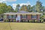 This charming one-story brick house on 309 20th Street in Butner features black shutters, white trim, and a cozy front porch with railings. It sits on a grassy lawn with neatly trimmed bushes and trees in the background.