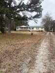 A single-story beige house with a green roof sits at the end of a gravel driveway on 1880 Long Mill Road in Youngsville, surrounded by brown grass, bare trees, and pine trees on a cloudy day.