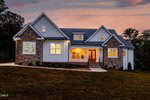 A two-story house with stone and white siding exterior is pictured at dusk. Large windows and front porch lights are illuminated. The house sits on a sloped, grassy yard with trees in the background.