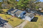 A single-story light blue house at 305 N Elm Street, Louisburg, with gray shingles, a front porch, and an attached shed sits amid a lawn and trees. A paved walkway and driveway are visible from above on a clear day.