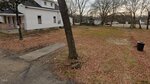 A vacant lot covered in brown leaves with a few scattered trees, next to a white house with a porch at 311 Piedmont Avenue, Oxford. A trash bin sits on the right side, with other houses and trees visible in the background.