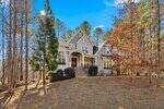 A large two-story house with beige siding at 3596 Bragg Valley Lane boasts an arched entryway, front porch, and multiple gables on a sloped, wooded Wake Forest lot with mature trees and sparse grass under a clear blue sky.
