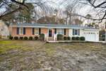Charming single-story brick house at 201 Drew Street, Louisburg, with dark shutters, white trim, a cozy front porch with railings, and an attached one-car garage. Sparse grass and bushes line the yard, with trees visible in the background.