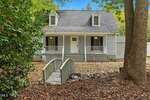 A gray two-story house at 109 Shearin Court, Youngsville, features dormer windows, a covered front porch with black shutters and a white door. A small wooden footbridge crosses rocky terrain, surrounded by trees and shrubs in the yard.