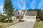 This two-story beige house with white trim at 95 Glen Oaks Drive in Youngsville features a two-car garage, a covered front porch, a large tree in the yard, and a wide driveway under a blue sky with wispy clouds.