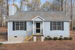 A small, light blue house with black shutters, gray shingle roof, central front door, and a wooden porch with steps sits at 164 Bridges Lane in Youngsville, surrounded by bare trees and brown ground cover.