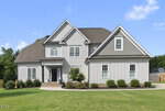 Charming two-story gray house with white trim at 210 Meadow Lake Drive, Youngsville, featuring a covered front porch, multiple gabled roofs, and a manicured lawn with small shrubs and trees on a sunny day.