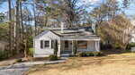 A small, single-story gray house with white trim at 1540 Cypress Drive, featuring a covered front porch, two chimneys, and a large front window, is nestled among trees and shrubbery in Henderson on a sunny day.