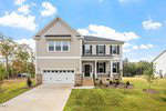 Charming two-story suburban house at 15 Guardian Street, Youngsville, featuring beige siding, white trim, stone accents, a covered front porch, two-car garage, manicured lawn, and a clear blue sky with scattered clouds.