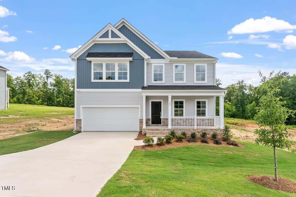 This charming two-story suburban home at 125 Cordoba Drive, Franklinton, features blue and white siding, an attached two-car garage, a welcoming front porch, manicured lawn, and young trees under a bright, clear sky.