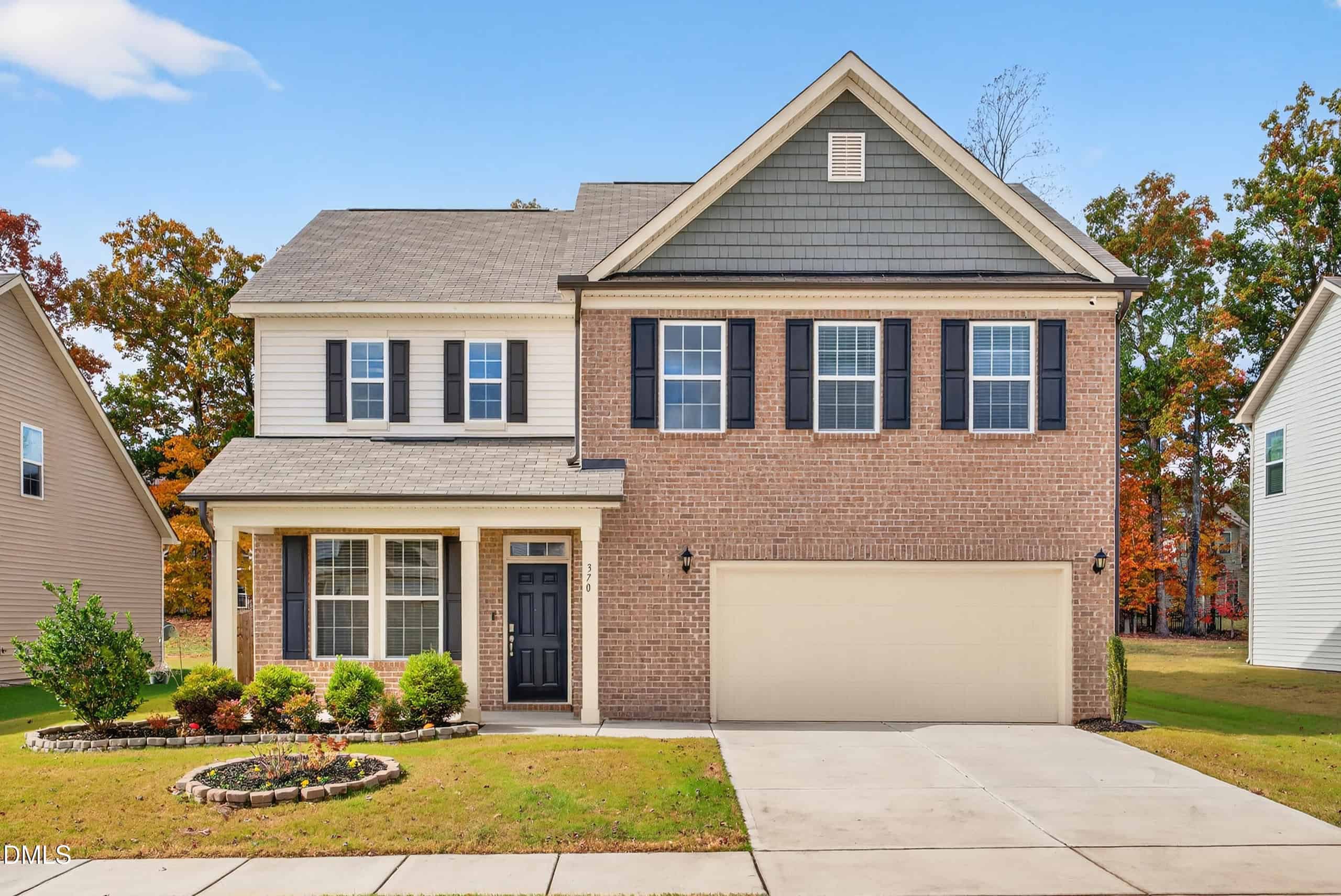 Charming two-story suburban house at 370 Hickory Run Lane, Youngsville, with brick and siding exterior, black shutters, double garage, and a cozy front porch. Features a neatly landscaped yard with a round garden bed and green shrubs.
