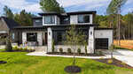 Modern two-story house at 95 Cherry Bark Drive, Youngsville, featuring a black and white exterior, large windows, front porch with seating, attached single-car garage, landscaped yard with young trees and neat grass. Forested backdrop completes the scene.