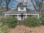 A small, white, single-story house at 847 Brantleytown Road in Zebulon features a central dormer window, a covered front porch with a red front door, overgrown bushes, fallen leaves, and leafless trees under a partly cloudy sky.