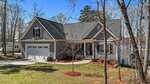 A gray, two-story house at 1013 Sagamore Drive, Louisburg, with a front porch, attached two-car garage, and gabled roof is nestled among trees and a grassy yard, with a lake visible in the background.