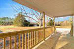 A wooden porch with a railing overlooks a spacious, grassy yard with scattered trees and a driveway. The sky is clear and sunny, casting shadows on the porch floor.