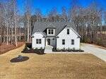 A newly built two-story white house with gray roofing sits on a large, freshly sodded lawn at 12874 Mallard Rock Drive in Zebulon. The driveway curves to the right, and the home is surrounded by a wooded area of tall, leafless trees.