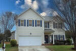 Charming two-story suburban house at 1300 Marbank Street, Wake Forest, with cream siding, blue shutters, and a two-car garage. White columns accent the front porch and a wreath adorns the door. Sparse trees frame a cloudy blue sky in the background.