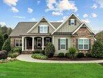 A modern suburban house at 15 Princeton Manor Drive, Youngsville, with gray siding and stone accents, features a covered front porch, manicured bushes, a green lawn, and a curved walkway under a blue sky with clouds.