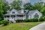 A light gray house with white trim at 180 Normandy Road, Louisburg features a large porch, dormer windows, and a gabled roof, surrounded by greenery and trees, with a sloped lawn and concrete driveway in front.