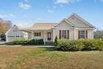 Charming single-story beige house at 160 Glenmore Circle in Youngsville with brown shutters, a covered front porch, and an attached two-car garage. The yard features grass and shrubs under a clear sky with a few clouds.