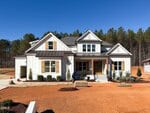 A modern farmhouse at 95 Spanish Oak Drive in Youngsville features white siding, wooden shutters, and a covered front porch overlooking a red dirt yard. Small shrubs dot the landscape, while tall trees frame the home beneath a clear blue sky.