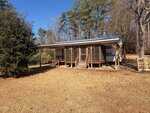 A small cabin with a covered porch sits in a grassy yard at 1561 Sagamore Drive, Louisburg, surrounded by trees. Steps lead up to the porch, supported by wooden posts, and pallets are stacked to the right of the building.