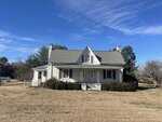 A small, white, two-story house with a front porch and dormer window sits in a large, grassy yard at 1116 New Bethel Church Road in Henderson. Bushes line the front of the house under a blue sky with scattered clouds.