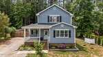 A charming two-story blue house with white trim on 225 Sacred Fire Road, Louisburg, featuring a covered front porch, surrounded by trees and greenery. A gravel driveway leads to the side and a small fenced area sits on the right.