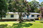 A white, single-story house at 202 Cooper Street in Louisburg features blue shutters and a brick foundation. Surrounded by green grass and pink-flowered trees, it has a ramp to the front door and sits beneath a bright, sunny sky.